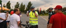 Na dworze, na wyłożonym kostką placu stoi mężczyzna w żółtej kamizelce, obok niego drugi mężczyzna trzyma rower.