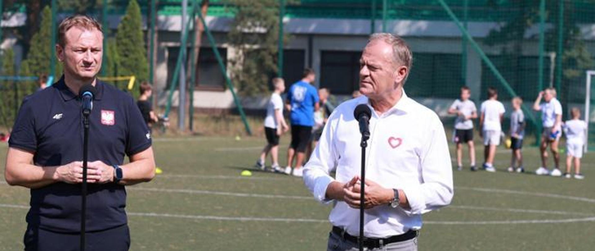 Ubrany na granatowo Minister Sportu i Turystyki Sławomir Nitras i Premier Donald Tusk w białej koszuli stoją przy mikrofonach na boisku. W tle dzieci grające w piłkę. Dalej siatka i zabudowania.