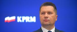 Minister Czarnek na tle niebieskiej ściany z logiem KPRM.