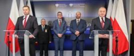 Z przodu przy dwóch mównicach z przezroczystego tworzywa stoją minister Czarnek i mężczyzna w garniturze, za nimi trzech mężczyzn, za nimi dalej ściana, po bokach z obu stron polskie flagi.