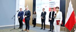 Na tle wielkiego ekranu z napisami NAWA stoi grupa ludzi, z frontu dwóch mężczyzn w garniturach przy mikrofonach na stojakach, z boku ubrana na biało kobieta mówi do trzymanego w ręku bezprzewodowego mikrofonu, za nią polska flaga.
