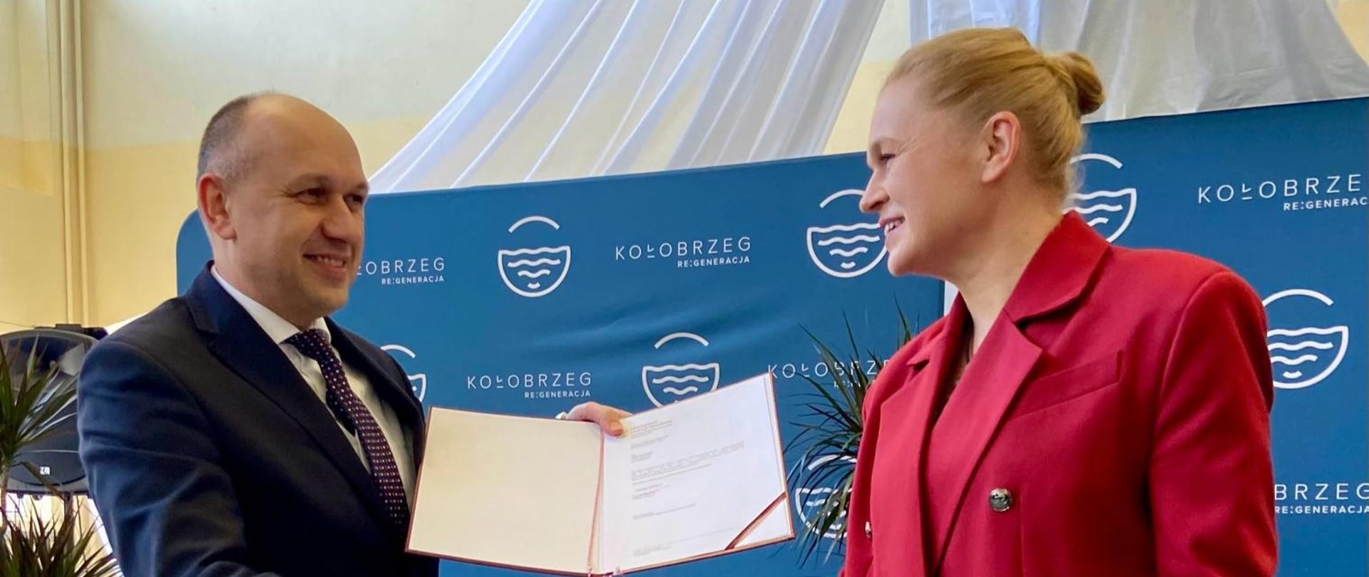 Minister Nowacka podaje rękę nowemu kuratorowi Pawłowi Palczyńskiemu, który trzyma otwarty akt powołania. W tle ścianka z napisem Kołobrzeg i zielone rośliny.