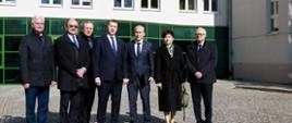 Na podwórku wyłożonym szarą kostką stoi sześciu mężczyzn w garniturach i kobieta w czarnym płaszczu, za nimi biały budynek z zielonymi przeszklonymi drzwiami i napisem Państwowy Instytut Weterynaryjny.