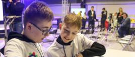Dwóch chłopców w białych koszulkach z napisami Liga Robotów siedzi przy białym stole, oglądają coś na tablecie.