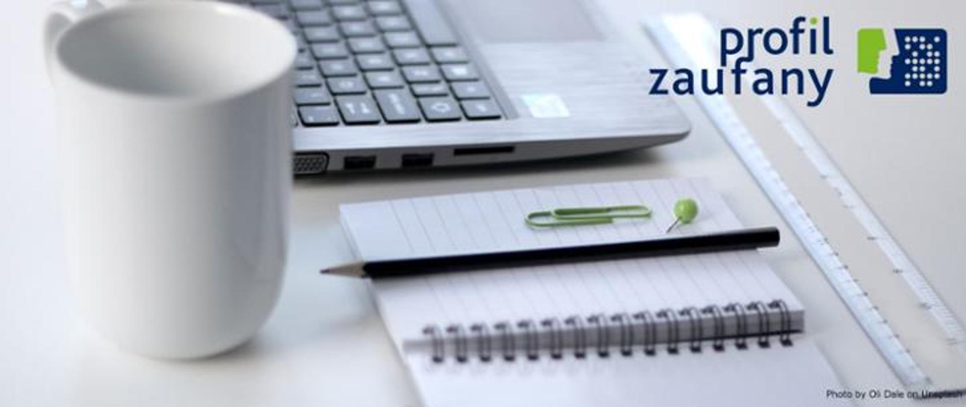 Grafika. Na biurku leżą laptop, notes i kubek. Obok napis: Profil Zaufany.