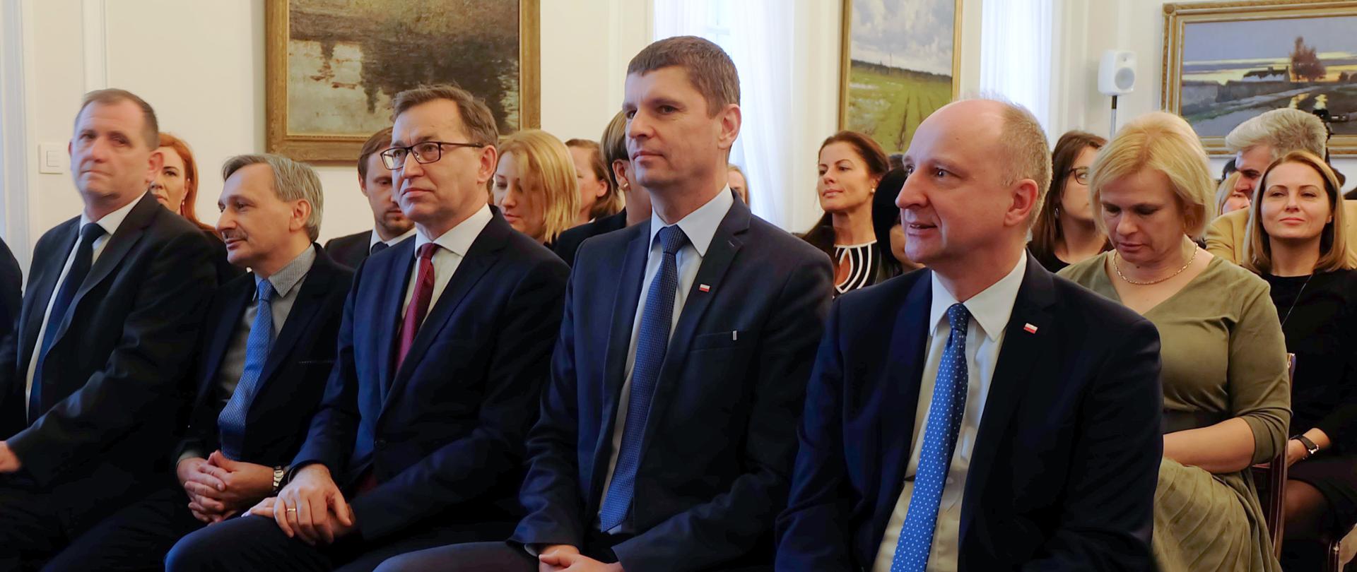 Minister edukacji siedzi w pierwszym rzędzie. Obok niego minister w kancelarii prezydenta i wiceminister edukacji. 