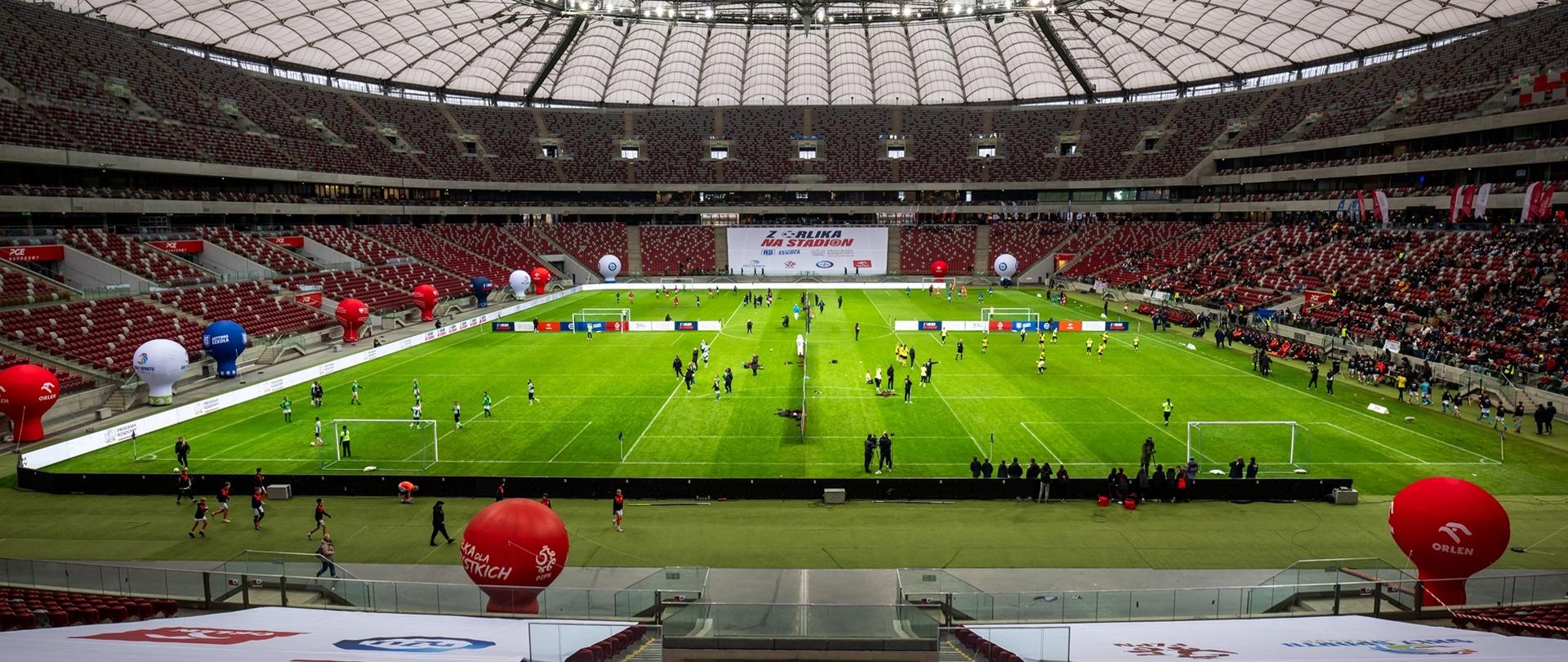 Stadion do gry w piłkę nożną. Na murawie widać kilka osób uprawiających sport 