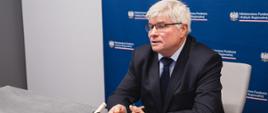 Wiceminister funduszy i polityki regionalnej Maciej Lasek siedzi przy stole z laptopem, za jego plecami baner z logotypem MFiPR