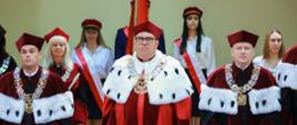 Grupa ludzi w ceremonialnych czerwonych strojach stoi za niebieskim stołem, za nimi żółtozielona ściana.