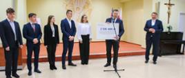Minister stoi na środku sali, trzyma w rękach duży symboliczny czek z napisem 2 126 400 zł, po jego lewej stronie stoi mężczyzna w garniturze, po prawej grupa elegancko ubranej młodzieży.