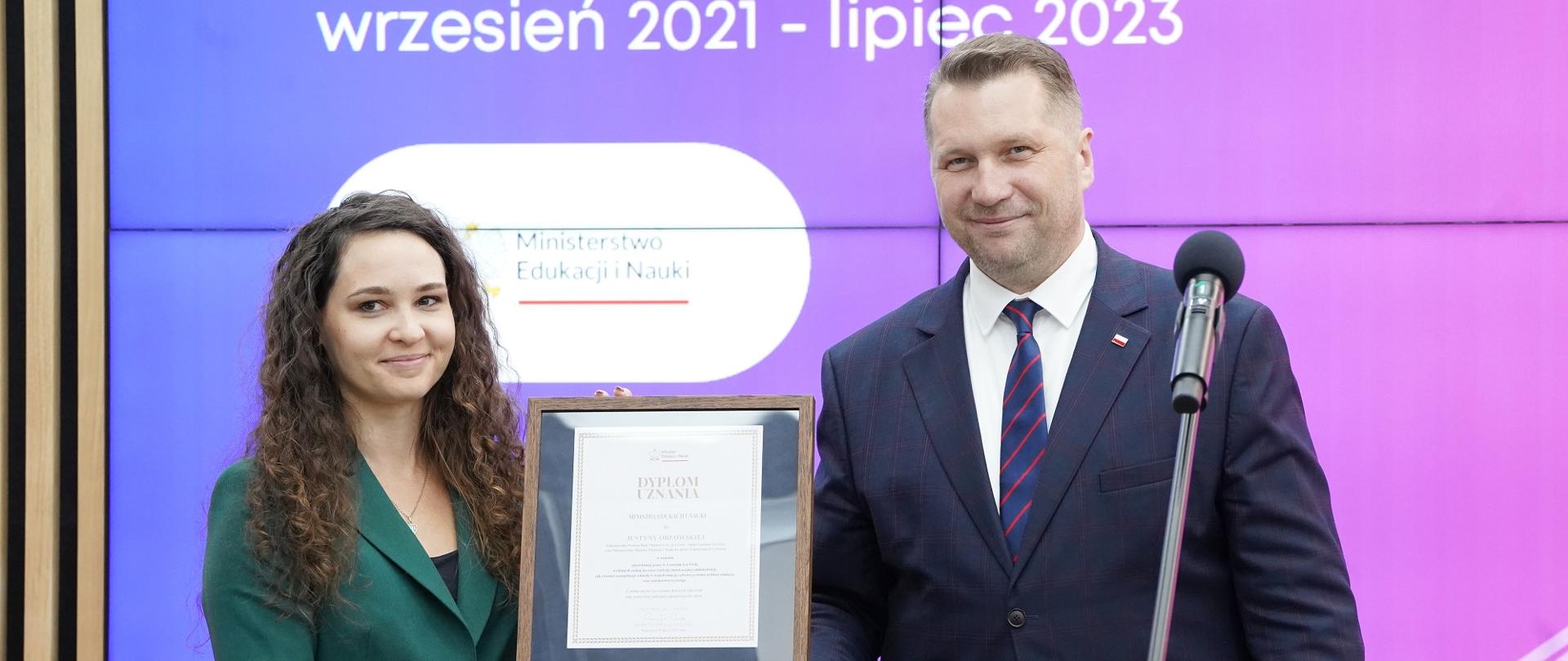 Minister Czarnek i kobieta w zielonej marynarce stoją obok siebie, razem trzymają dokument w ramkach.