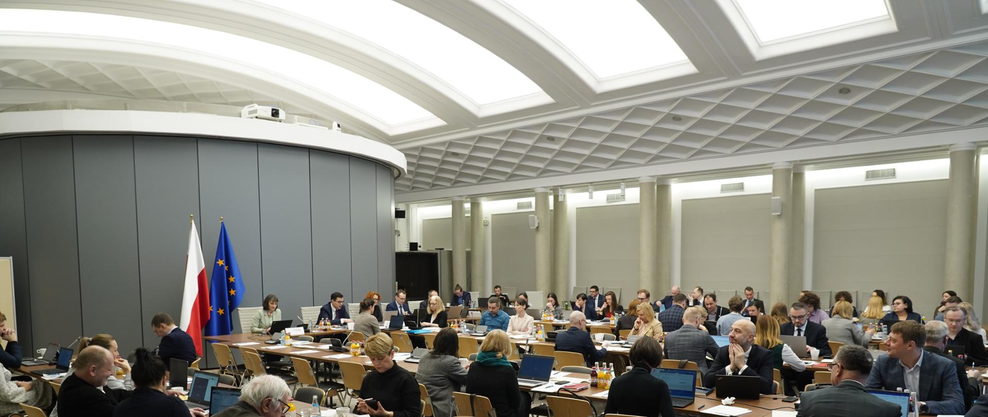 Kilkadziesiąt osób siedzi na sali przy długich stołach. 
