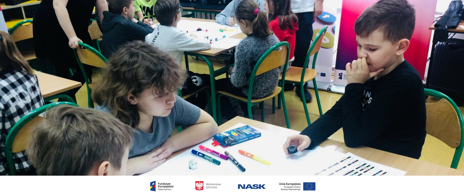 Uczniowie siedzący przy stoliku. Na stoliku kolorowe pisaki.
