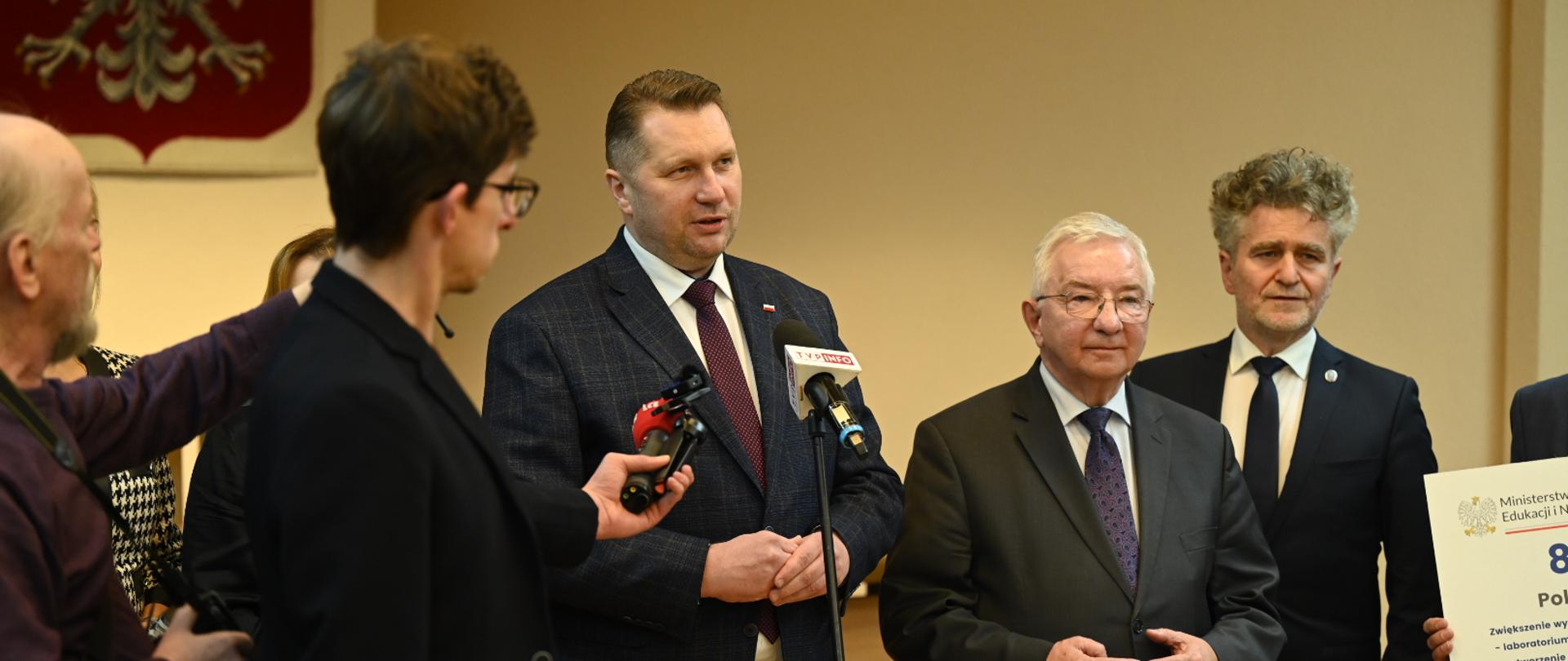 Grupa osób wokół ministra Przemysław Czarnka. Dziennikarze trzymają mikrofonu. 