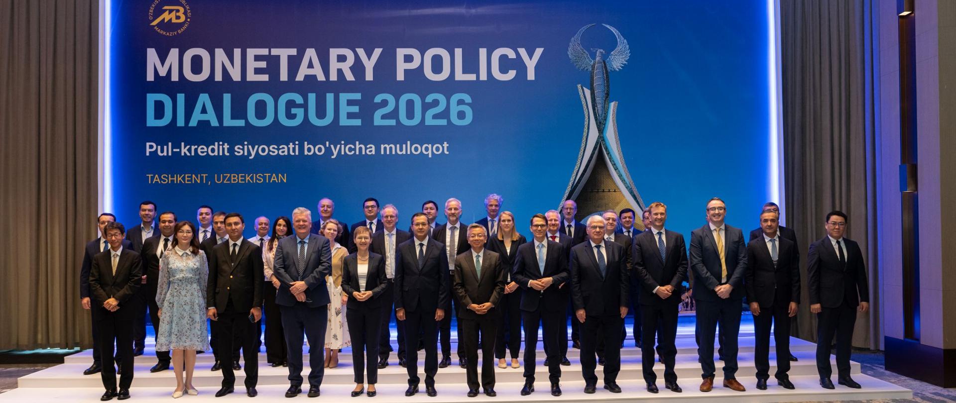Grupa uczestników wydarzenia Monetary Policy Dialogue 2026 pozuje do wspólnego zdjęcia na scenie przed dużym ekranem z nazwą konferencji i lokalizacją w Taszkencie w Uzbekistanie.