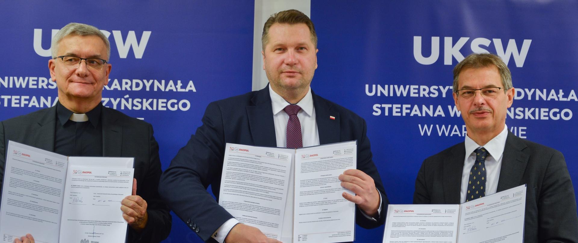 Minister Czarnek i dwóch mężczyzn w garniturach stoją pod ścianą z napisami UKSW, każdy trzyma otwarty dokument.