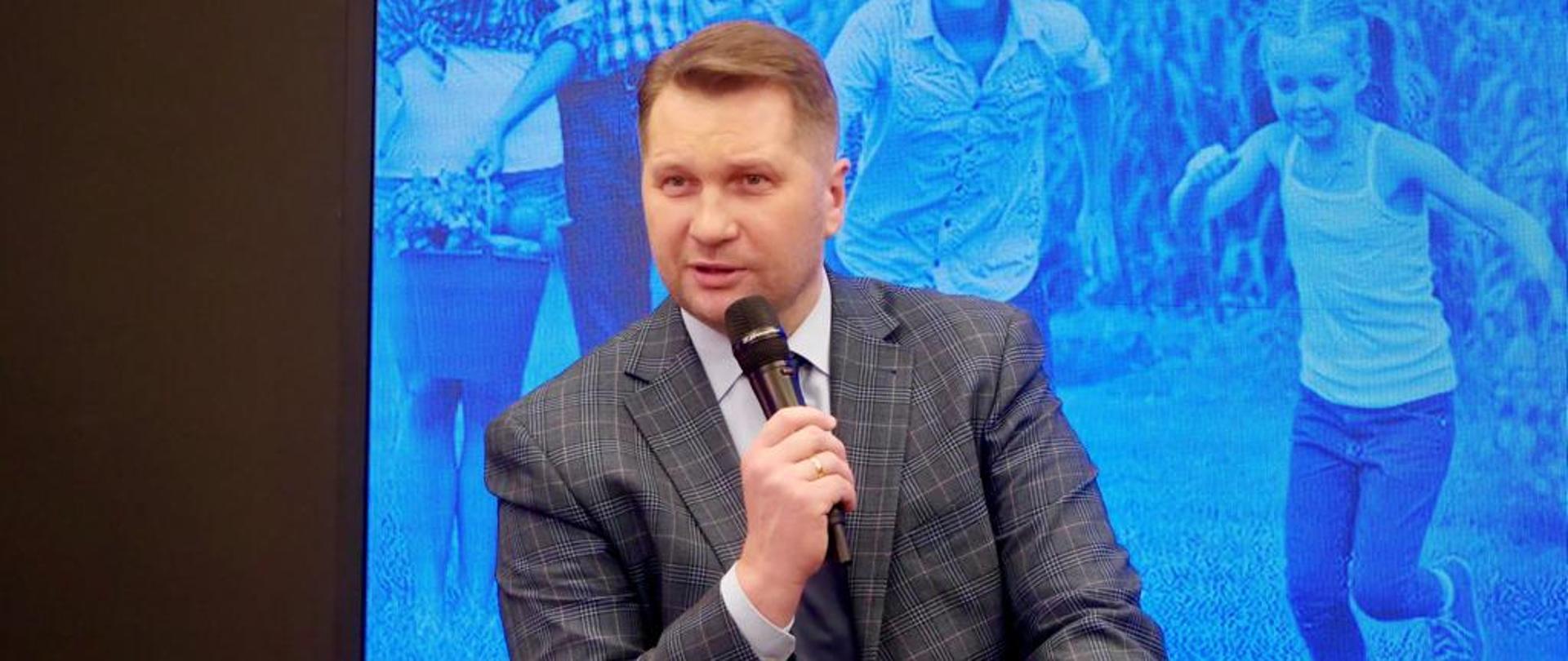 Debata z okazji Dnia Praw Rodziny minister edukacji i Nauki Przemysław Czarnek przemawia na siedząco do uczestników. W ręku trzyma mikrofon. W tle niebieski ekran na którym biegają rodziny z dziećmi