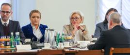 Przy stole siedzi kilka osób, wśród nich wiceminister Machałek w białym ubraniu, za nimi biała ściana i białe zasłony na oknach.