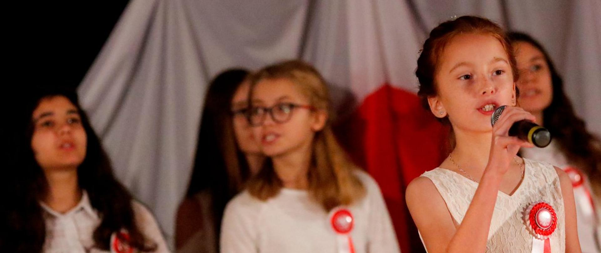 Dzieci śpiewają hymn Polski, w tle ciało-czerwona flaga.