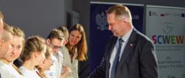 Minister Czarnek stoi naprzeciwko szeregu dzieci ubranych w białe koszulki, podaje rękę dziewczynce, za nimi baner z napisem SCWEW.