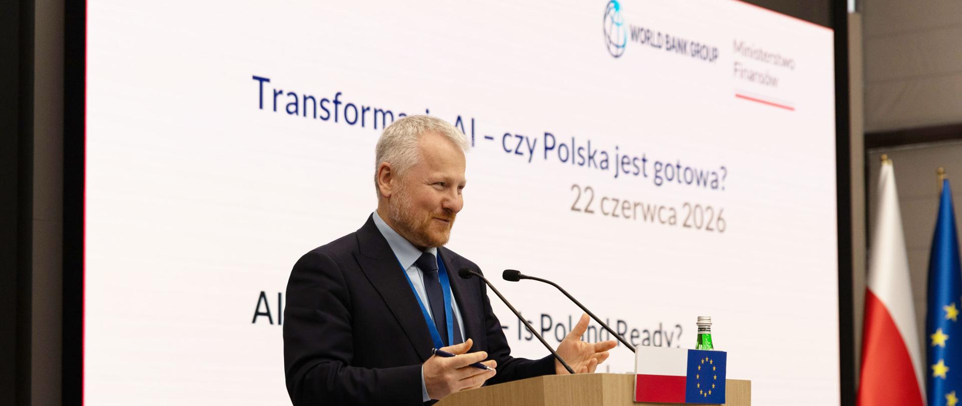 Wiceminister finansów Jurand Drop przy mównicy i przemawia podczas wydarzenia dotyczącego sztucznej inteligencji, na tle ekranu z napisem „Transforming AI – czy Polska jest gotowa?” oraz flag Polski i Unii Europejskiej.
