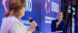Minister Barbara Nowacka na scenie konferencyjnej, słuchająca pytania lub wypowiedzi innej osoby (widocznej nieostro na pierwszym planie). Ministra uśmiecha się lekko, trzymając mikrofon.