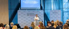 Szeroki plan na salę konferencyjną. Kobieta w białym garniturze przemawia przy mównicy na tle ścianki z logo Ministerstwa Edukacji Narodowej. Nad nią znajduje się duży ekran z napisem: „KONFERENCJA DIAGNOZA MŁODZIEŻY 2026. OD OCENY SYTUACJI DO STRATEGII WSPARCIA MŁODZIEŻY I MŁODYCH DOROSŁYCH”. Na pierwszym planie widoczne są liczne rzędy siedzących uczestników, słuchających wystąpienia w nowoczesnym, przeszklonym wnętrzu.
