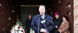 Minister Czarnek stoi na dworze i mówi do trzech mikrofonów na stojakach, za nim ciemne pomieszczenie, obok biało-czerwony wieniec i kilka osób.