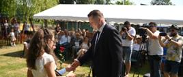 Dziewczynka podaje ministrowi Czarnkowi kwiat, stoją na trawie, za nimi namiotowy daszek pod którym siedzi grupa dzieci.