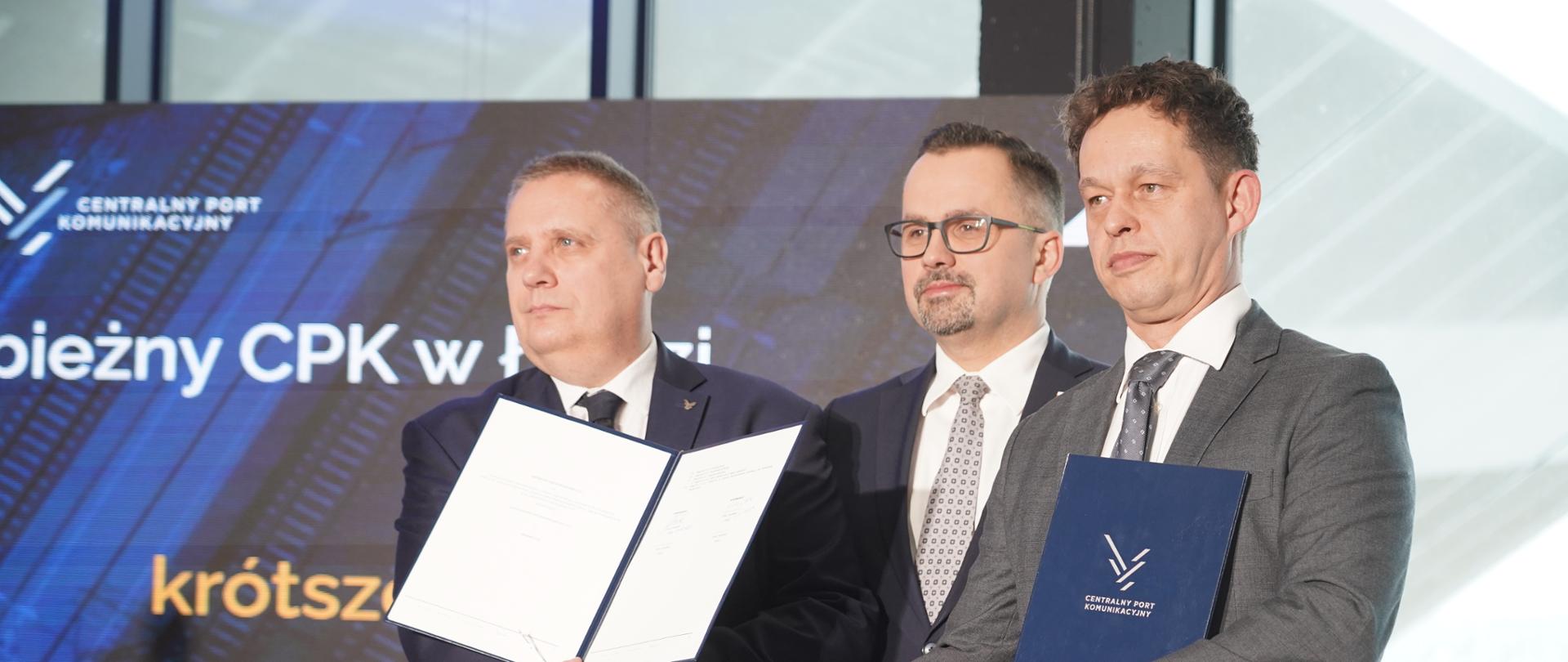 Trzech mężczyzn stoi obok siebie. Dwóch z nich podaje sobie ręce. Trzymają też podpisane uroczyście umowy. Pośrodku stoi minister Marcin Horała.