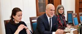 Przy drewnianym stole siedzi uśmiechnięty mężczyzna w garniturze, po obu stronach dwie kobiety - ubrana na czarno i w marynarce w czerwono-czarną kratę, za nimi biała ściana i drewniana szafka z książkami.