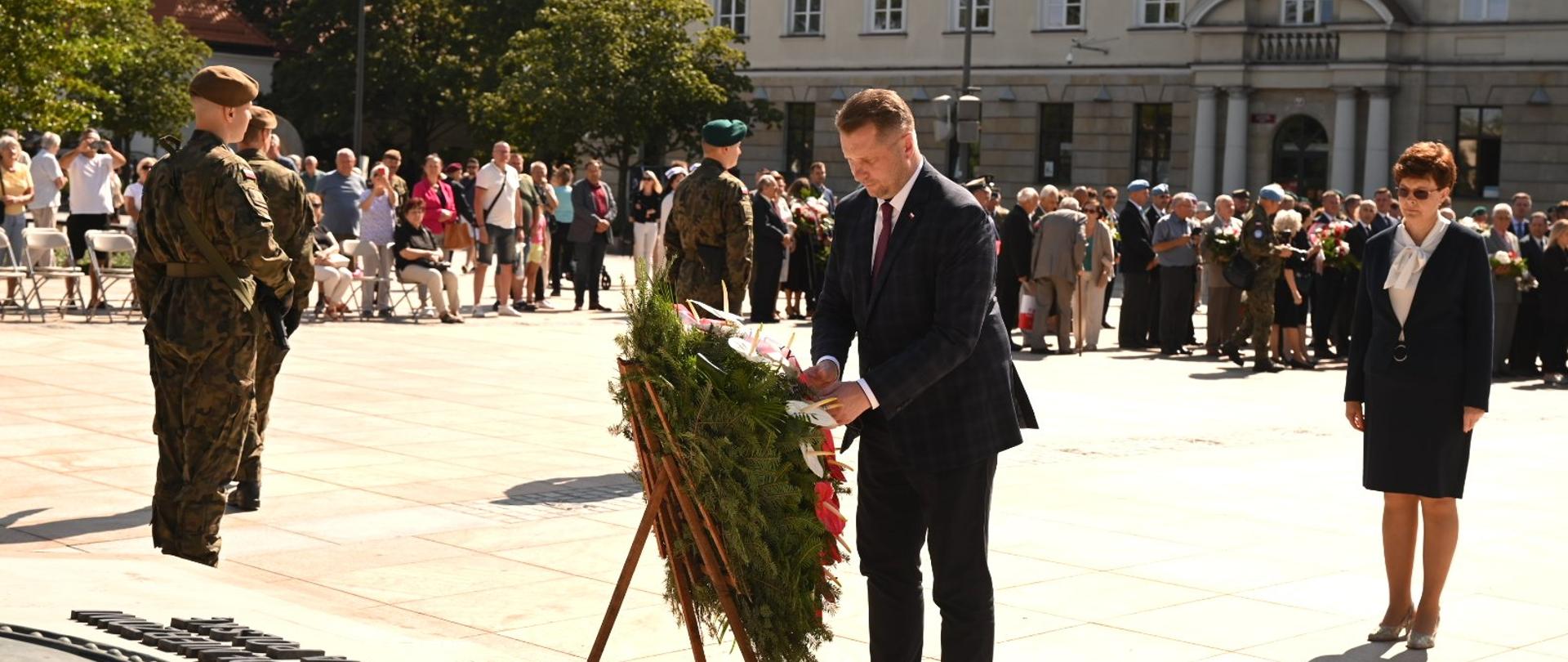 W ostrym słońcu na wyłożonym kamiennymi płytami placu minister Czarnek ustawia wieniec na drewnianej podstawce przed pomnikiem.