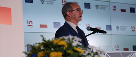 minister Jerzy Kwieciński mówi do mikrofonu, za nim symbole 15 lat Polski w UE