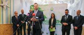 Minister Czarnek stoi i mówi do kilku mikrofonów na stojakach, za nim pięciu mężczyzn w garniturach i kobieta w marynarce w czarno-żółtą kratkę.