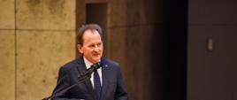 Wiceminister Bernacki stoi przy mównicy, obok kwiaty 