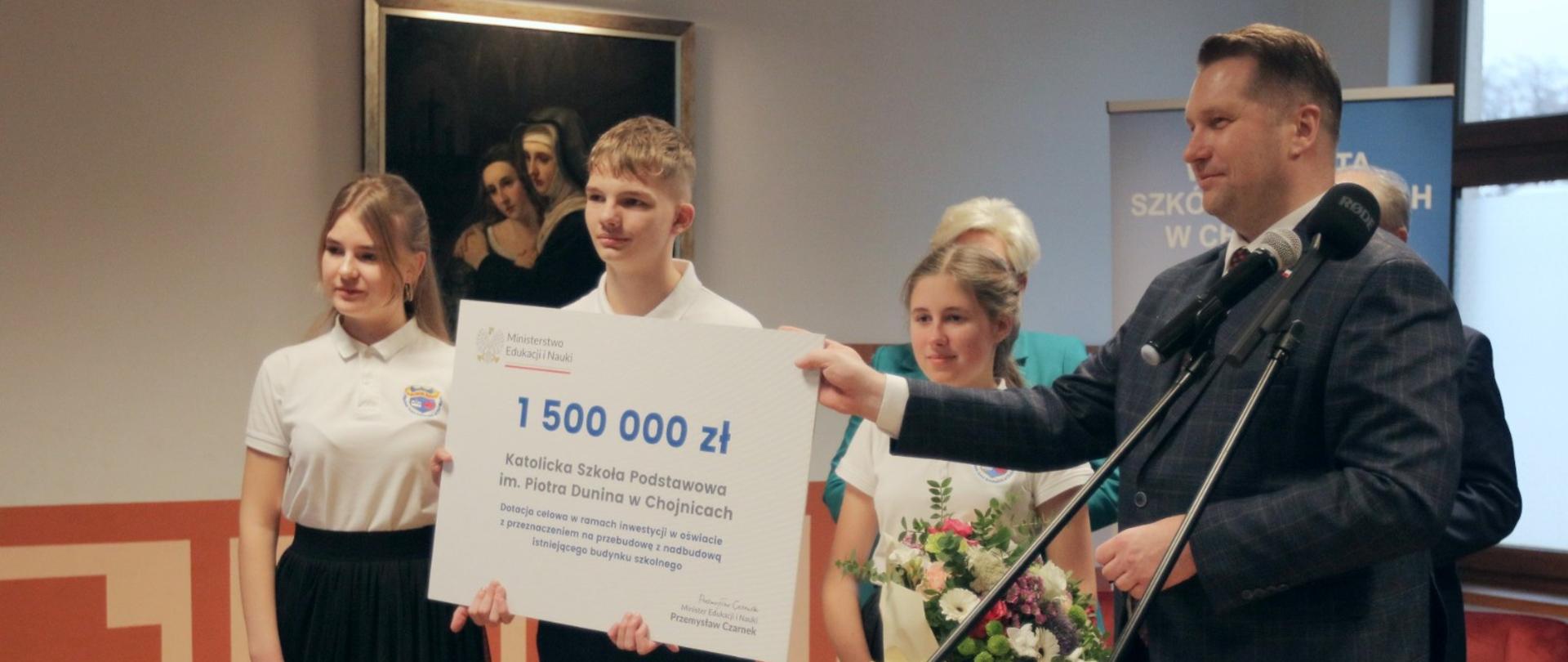 Minister Czarnek stoi przed dwoma mikrofonami na stojakach, obok niego dziewczyna z bukietem kwiatów, uczeń trzymający wielki symboliczny czek z napisem 1 500 000 zł, i jeszcze jedna uczennica. W tle na ścianie obraz.