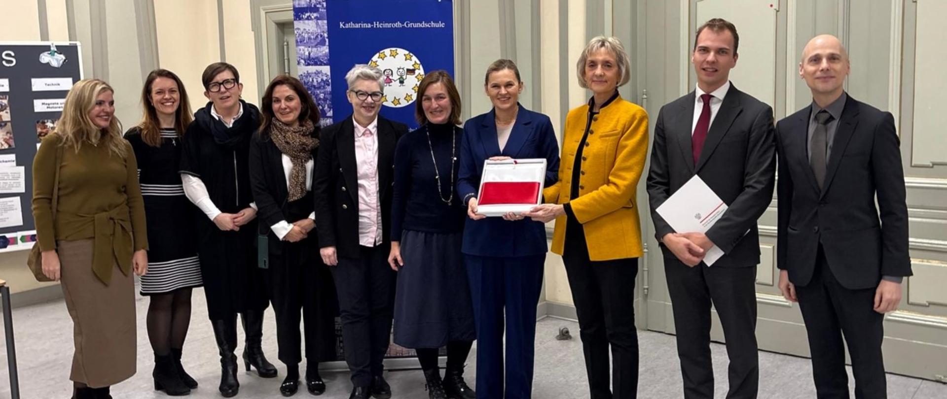 Zdjęcie grupowe dziewięciu osób, w tym Minister Edukacji Barbary Nowackiej (w granatowym garniturze, w centrum) oraz kobiety w żółtej marynarce. Minister Nowacka i kobieta w żółtej marynarce trzymają razem czerwonobiały dokument lub folder. W tle widoczny jest baner z napisem "Katharina-Heinroth-Grundschule".