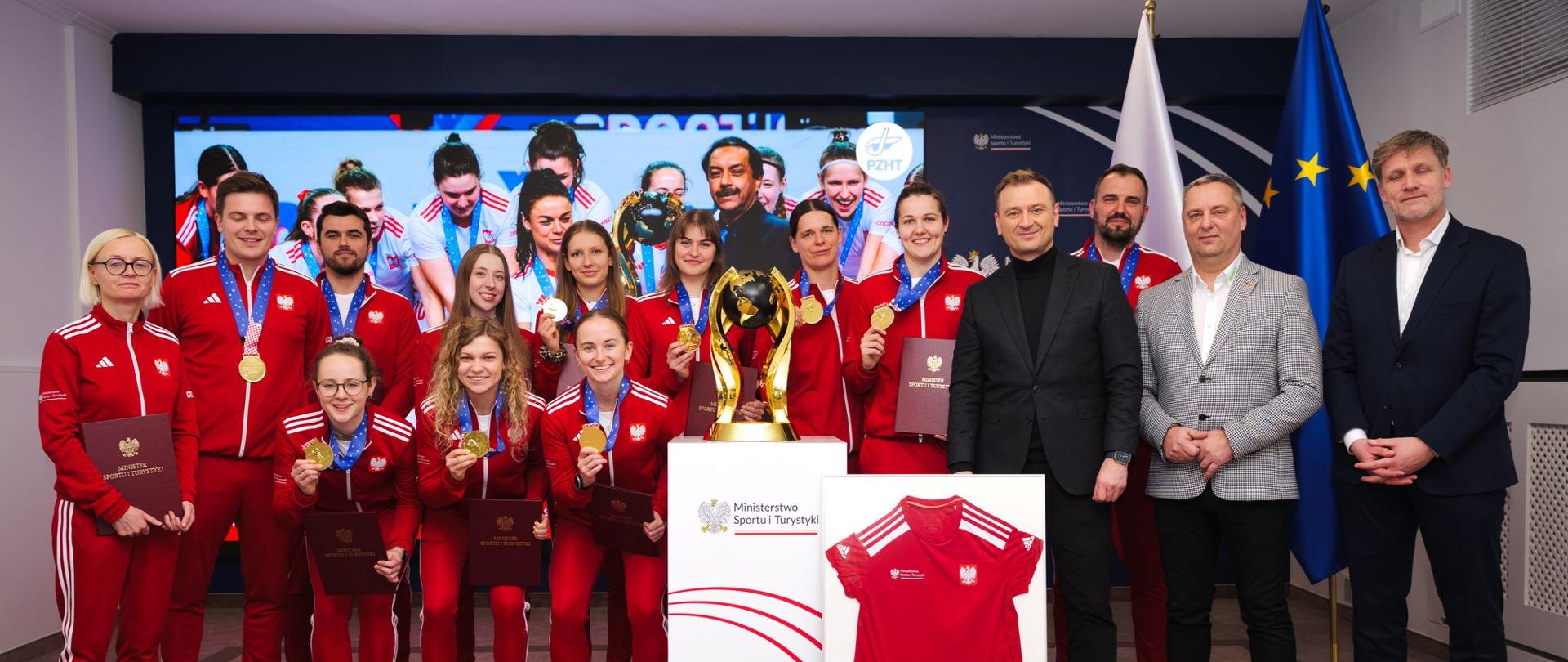Drużyna halowych mistrzyń świata w hokeju na trawie z ministrem sportu na tle ekranu ze zdjęciami