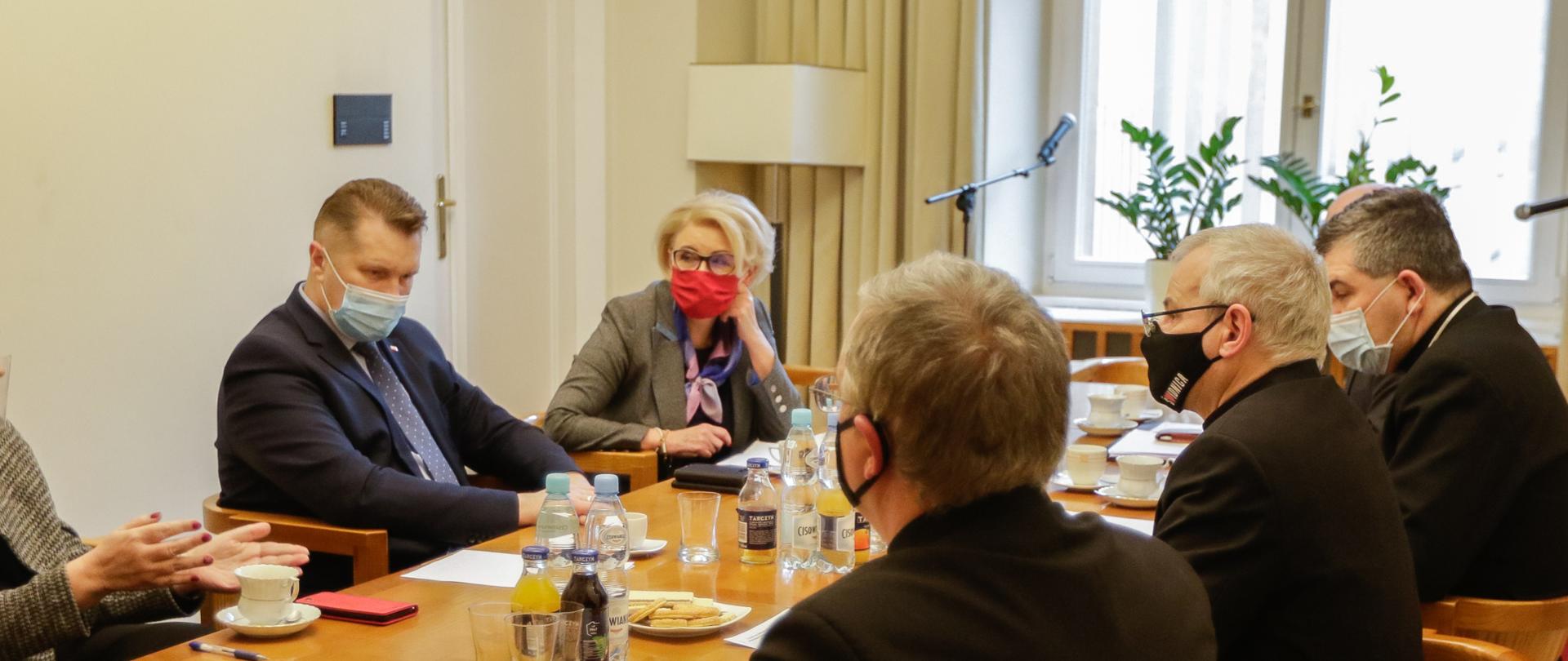 Przy podłużonym stole siedzą uczestnicy spotkania. Po jeden stronie przedstwiciele Ministerstwa Edukacji i Nauki minister Przemysław Czarnek oraz wiceminister Marzena Machałek. Po drugiej stronie trzej biskupi. Na stole stoją filiżanki, napoje, ciastka. Wszyscy mają notatki przed sobą na stole. 