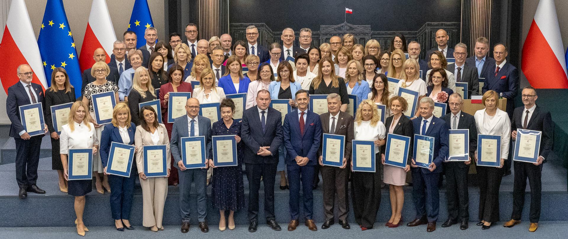 Duża grupa osób stoi na scenie i prezentuje oprawione certyfikaty podczas oficjalnej uroczystości, na tle flag Polski i Unii Europejskiej oraz ściany z logo instytucji