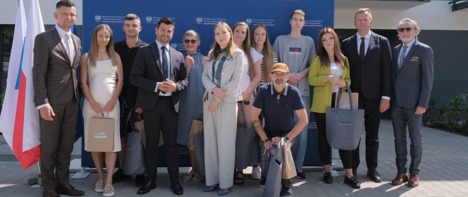 Grupa osób stoi obok siebie przed blokiem. Wśród nich jest minister Katarzyna Pełczyńska-Nałęcz.