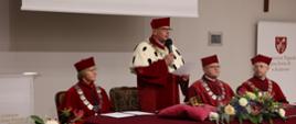 Przy nakrytym czerwonym materiałem stole siedzą trzy osoby w czerwonych ceremonialnych strojach, pośrodku stoi mężczyzna w takim samym stroju z gronostajowym kołnierzem i mówi do trzymanego w ręku mikrofonu.