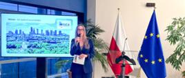 Osoba w garniturze prezentuje slajd o Warszawie na dużym ekranie, stojąc obok flag Polski i Unii Europejskiej.