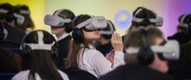 W sali z kolorowymi ścianami siedzi wielu młodych ludzi, na głowach mają gogle do wirtualnej rzeczywistości.