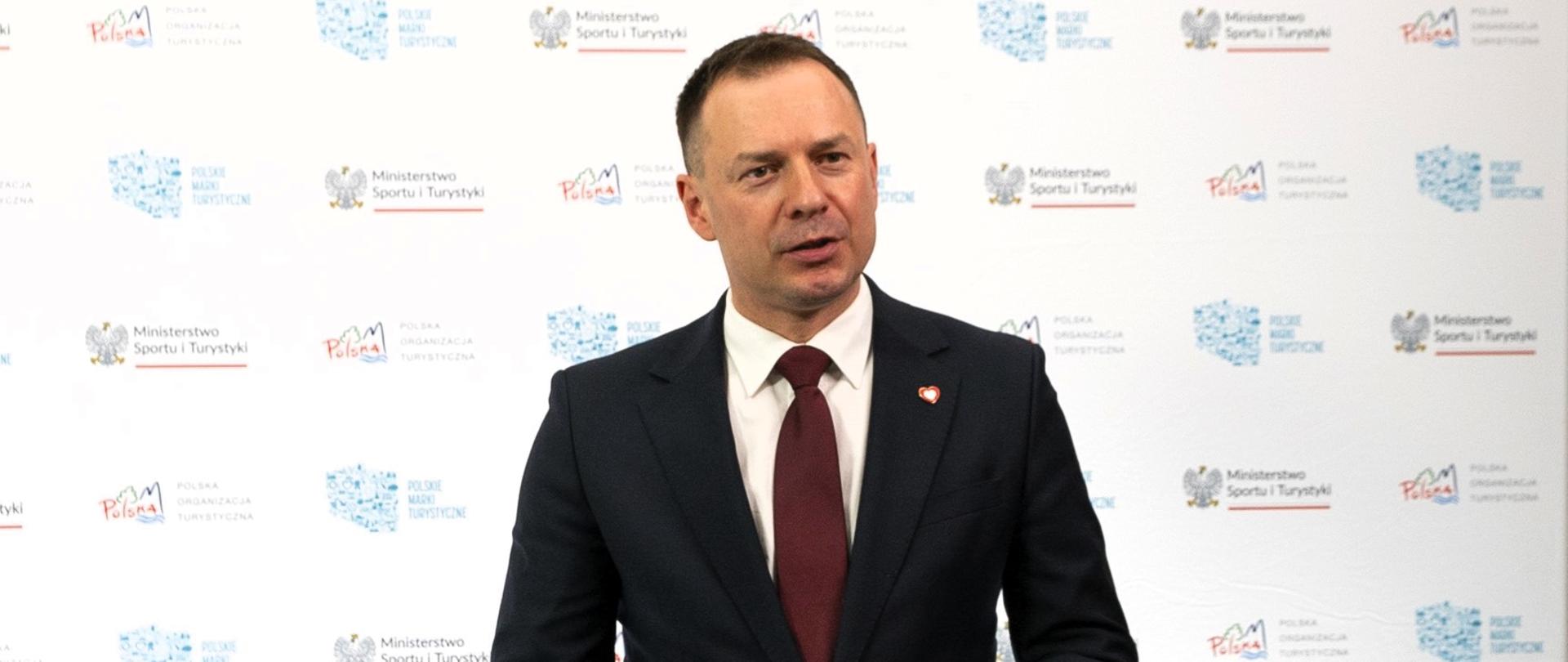 Zdjęcie przedstawiającego wiceministra sportu i turystyki Piotra Borysa, ubranego w ciemny garnitur z czerwonym krawatem. Wiceminister mówi do stojącego przed nim mikrofonu, a w tle stoi biała ścianka z logotypami Ministerstwa Sportu i Turystyki, Polskiej Organizacji Turystycznej oraz Polskiej Marki Turystycznej.