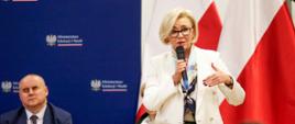 Wiceminister Machałek w białym ubraniu stoi i mówi do mikrofonu, za nią granatowa ściana i dwie polskie flagi.