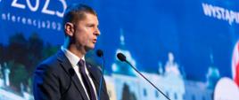 Wiceminister Piontkowski stoi przy mównicy i mówi do dwóch mikrofonów.