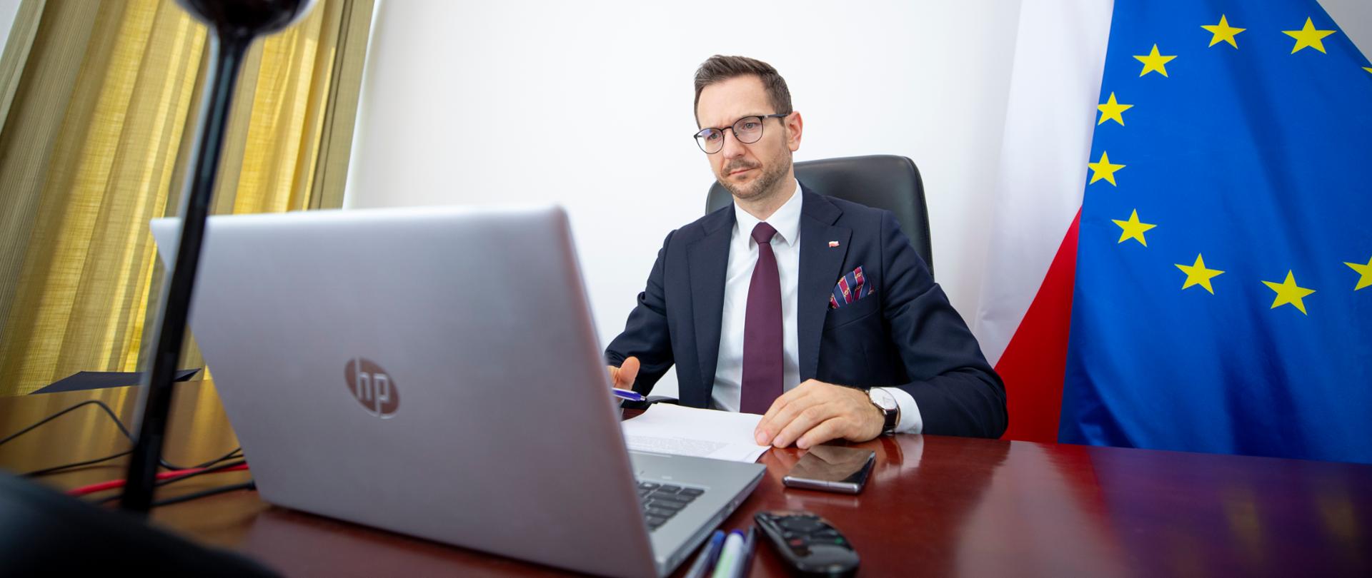 Wiceminister Waldemar Buda siedzi przy biurku, na którym stoi laptop i lampka. Za nim flagi PL i UE. Na ścianie wisi godło Polski.