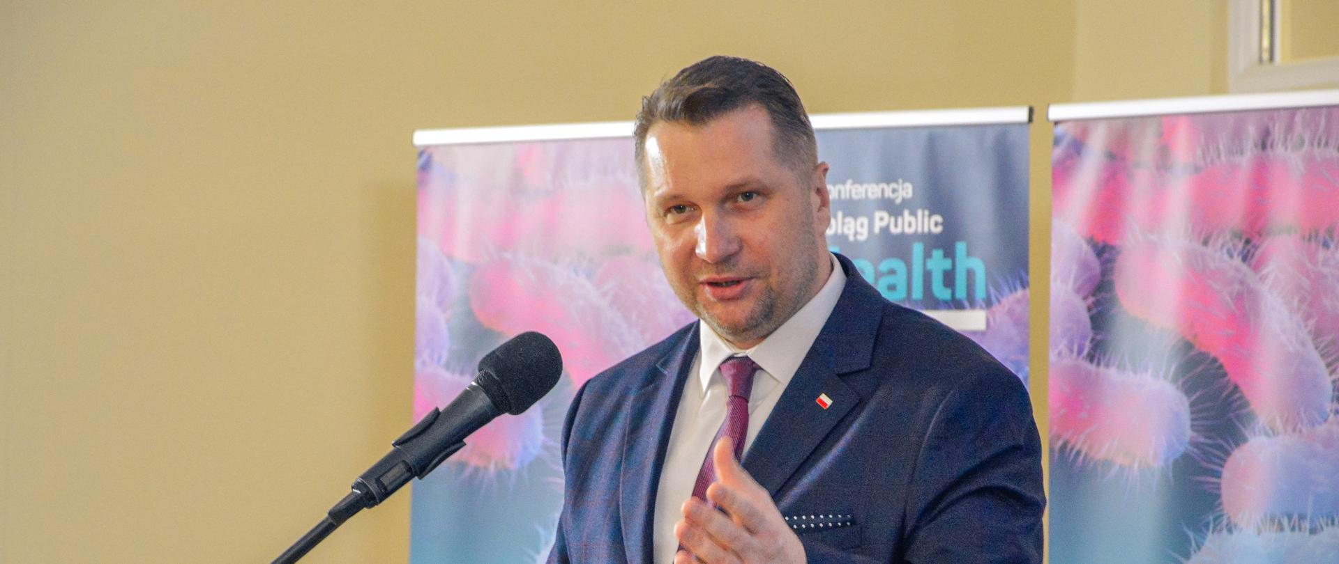 Minister Czarnek stoi przy mównicy i mówi do mikrofonu na stojaku, za nim przed żółtą ścianą dwa rozwinięte niebiesko-różowe banery.