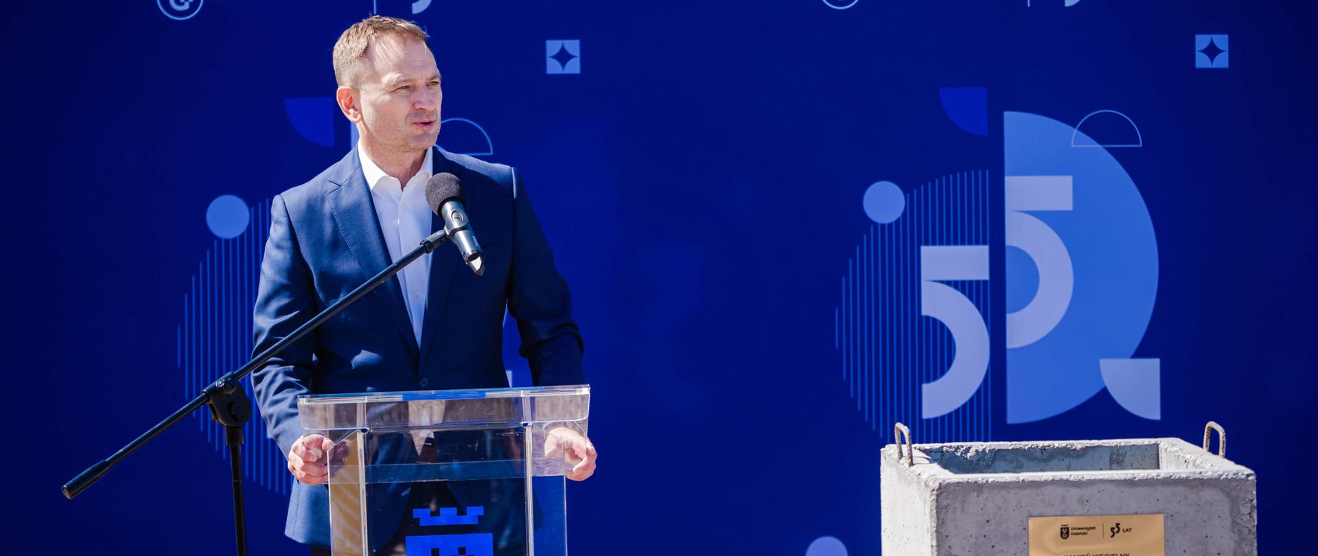 Minister Nitras stoi za mównica z logotypem UG i mówi do mikrofonu. W tle granatowa ścianka z logotypami Uniwersytetu Gdańskiego. Z prawej strony zdjęcia kamień węgielny z przymocowaną złotą tabliczką z napisem: "kamień węgielny pod budowę Centrum Sportowego Uniwersytetu Gdańskiego. Gdańsk, 28 kwietnia 2025 r.".